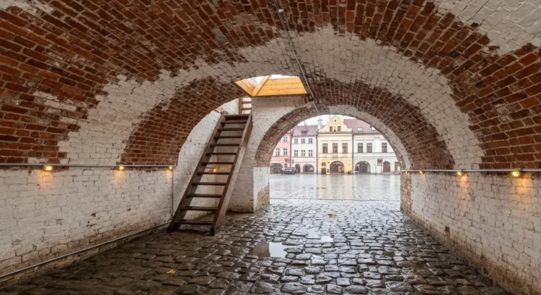Ceglane sklepienie podziemnej piwnicy pod rzeszowskim Rynkiem z drewnianymi schodami i widokiem na mokry od deszczu bruk